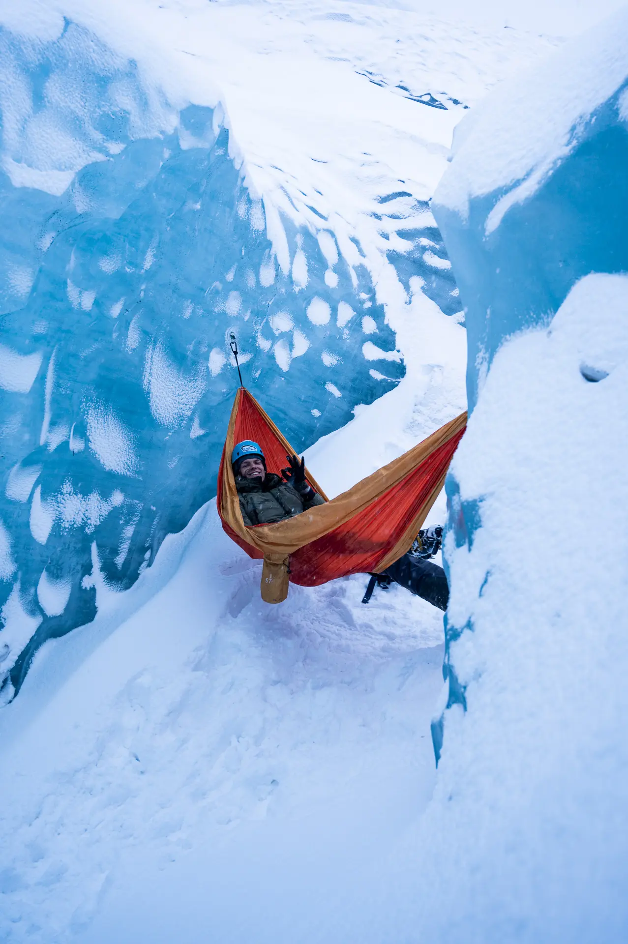 Man lays in a bright orange hammock strung up between two vivid blue ice walls, above a snow covered ground.