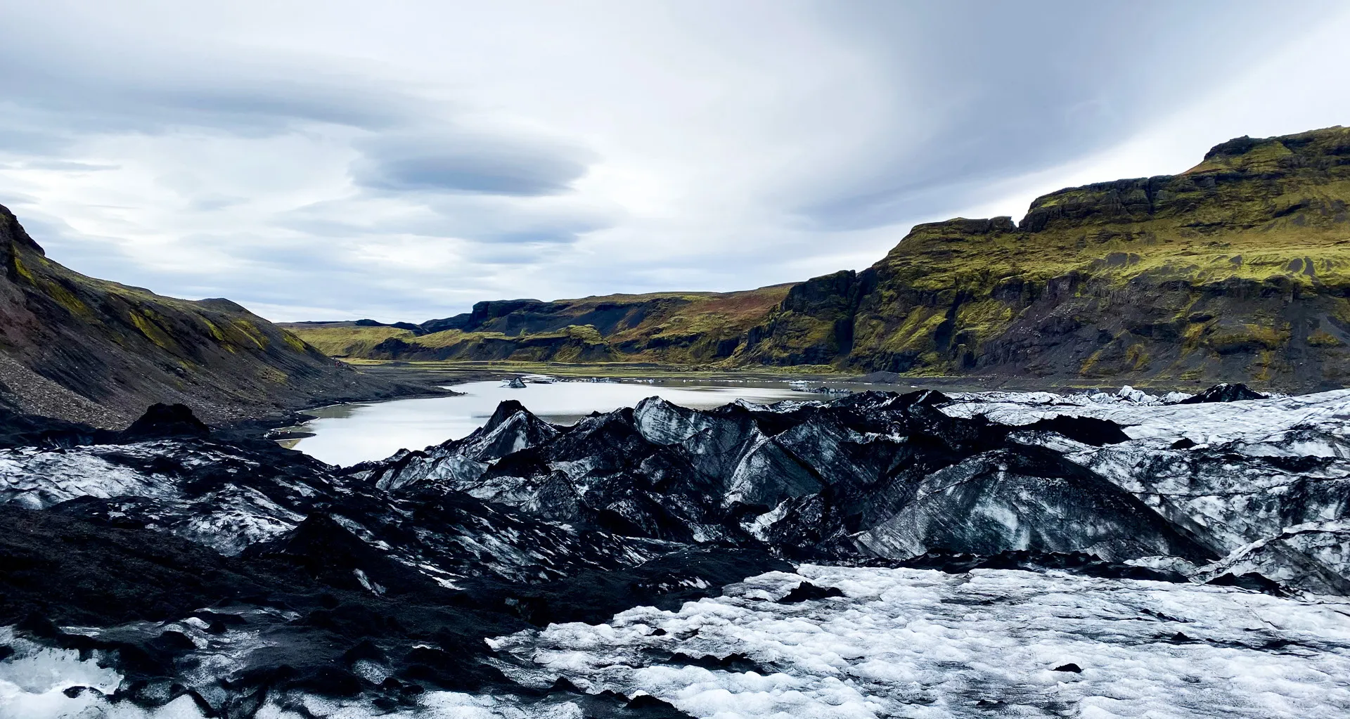 Mysterious clouds in overcast skies loom over black ash- covered ice on Sólheimajökull glacier with rugged, bright green mossy cliffs in the background.