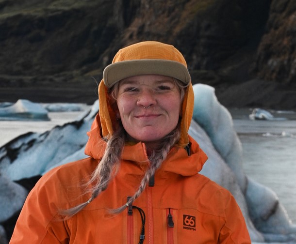 A woman in an orange jacket with braids poses on the glacier with a lagoon and ice peak in the background.